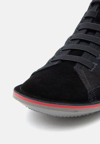 Schwarzer Wildleder-Sneaker mit strukturierter Oberseite, flachen Schnürsenkeln und abgerundeter Spitze. Die Sohle ist grau mit einem roten Akzentstreifen.