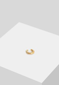 Anillo de oro con un diseño circular abierto, textura suave y acabado pulido, colocado sobre una superficie blanca con una ligera sombra.