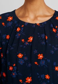 Granatowa sukienka w kwiaty z pomarańczowymi i niebieskimi kwiatami, z okrągłym dekoltem, plisami z przodu i małym czarnym guzikiem jako detalem.
