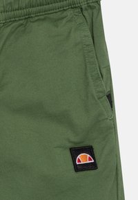 Groene shorts met een elastische tailleband, voorzien van een zijzak en een zwart geborduurd logo met een zonsondergangontwerp. Soepele stoftextuur.