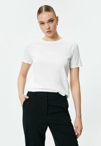 Witte katoenen T-shirt met korte mouwen, met een subtiele "ATTITUDE" tekstontwerp op de voorkant. Gecombineerd met zwarte getailleerde broek.