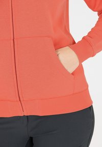 Koraalrode hoodie met rits, gemaakt van zachte stof, met ribgebreide manchetten en zoom, en voorzijde met kangoeroezakken. Gladde textuur, casual pasvorm.