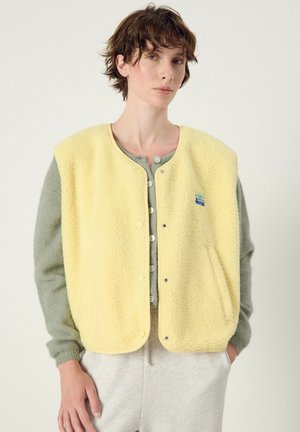 Person wearing a light yellow fleece vest over a gray buttoned cardigan and light gray pants, standing with one hand in pocket.