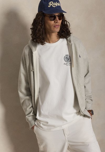 Mann mit langen lockigen Haaren, der eine dunkelblaue "Ralph"-Kappe, eine dunkle Sonnenbrille, einen beigen Kapuzenpullover mit Reißverschluss, ein weißes Ralph Lauren T-Shirt und weiße Hosen trägt.