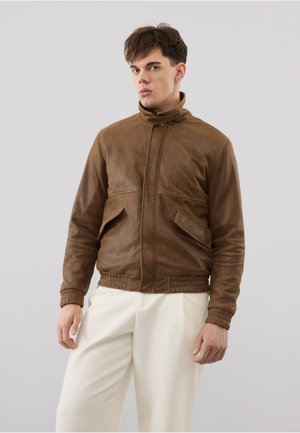 Junger Mann trägt eine braune Lederjacke mit vorderen Klapptaschen und weiße Hose, steht vor einem schlichten grauen Hintergrund.