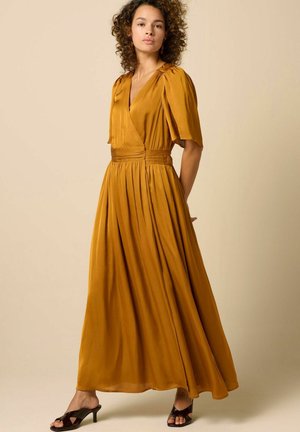 Frau mit lockigem Haar, die ein senfgelbes, kurzärmliges, plissiertes Maxikleid und schwarze Sandalen mit Absatz vor einem schlichten Hintergrund trägt.