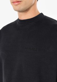 Sudadera negra con cuello redondo acanalado, que presenta un sutil logotipo embosado "PEGADOR" en el pecho. Textura suave, diseño minimalista.