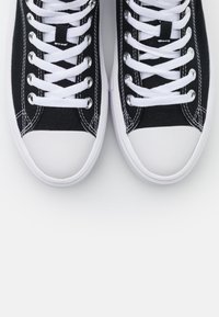 Paire de baskets en toile noire avec embouts en caoutchouc blanc et lacets blancs, vue de dessus sur fond blanc.