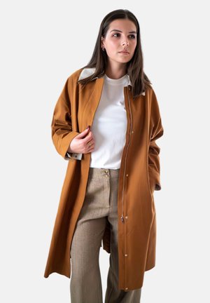 Bruine waterdichte jas met een witte kraag, voorrits en drukknoopjes. Draagt over een wit shirt en beige textuurbroek.