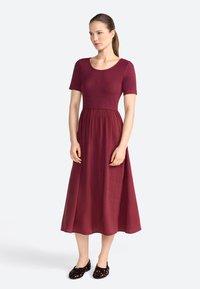 Robe maxi bordeaux à manches courtes, corsage ajusté et jupe fluide. Texture lisse, détail brodé sur la poitrine, et chaussures plates.