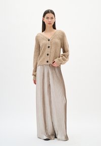 Kobieta w beżowym swetrze z guzikami i błyszczących, szerokich, plisowanych spodniach, stojąca z jedną ręką w kieszeni na białym tle.