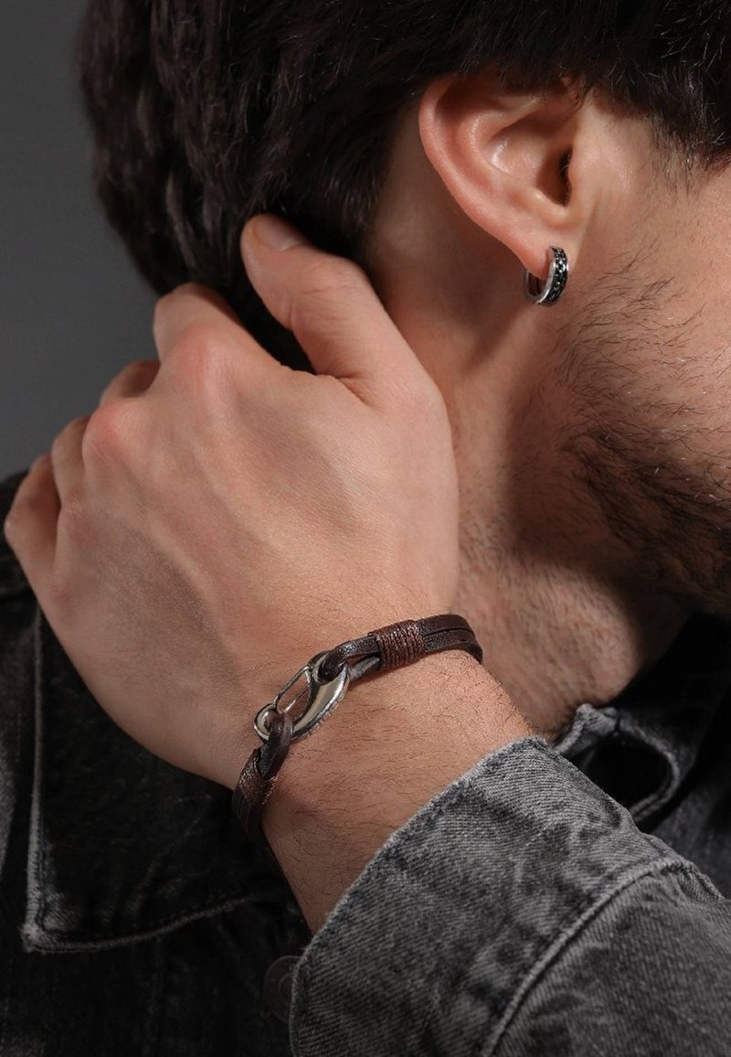 Braccialetto in pelle marrone con hardware in argento e accenti intrecciati, indossato su una mano che tocca un collo. Tessuto scuro e strutturato sullo sfondo.