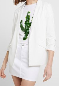 White blazer with rolled sleeves over a white t-shirt featuring a green cactus design, paired with a white denim skirt.