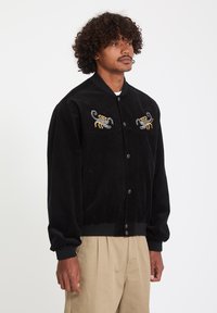 Homme portant une veste noire boutonnée avec des scorpions brodés jaunes et gris sur la poitrine, pantalon beige, debout devant un fond uni.