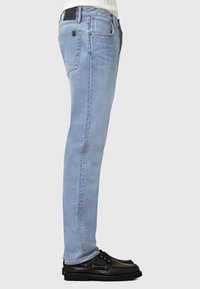 Jeans in denim azzurro chiaro con vestibilità slim, dotati di un'etichetta nera sulla vita, cuciture sottili e texture vissuta. Scarpe nere completano il look.