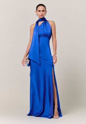 Kobieta w bezrękawnikowej sukni z satyny w królewskim niebieskim kolorze z wysokim rozcięciem, srebrne szpilki, pasująca kopertówka, geometryczne kolczyki i bransoletka na neutralnym tle.