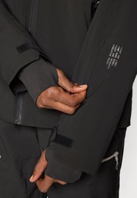 Zwarte technische jas met een gladde textuur, voorzien van duimopeningen, klittenbandsluitingen aan de mouwen en bedrukte tekst op de mouw. Ritsdetails zichtbaar.