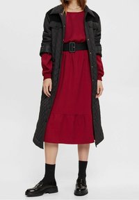 Manteau long matelassé noir sur une robe rouge à ourlet en cascades, avec une taille ceinturée, porté avec des chaussettes noires et des chaussures brillantes.