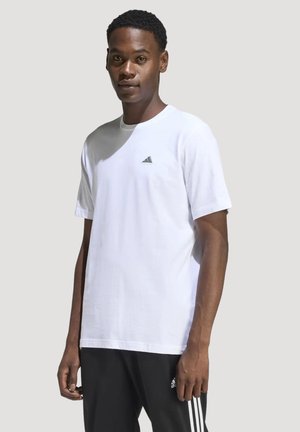 Junger Mann, der ein weißes Adidas-T-Shirt und schwarze Adidas-Trainingshose trägt, steht vor einem schlichten hellen Hintergrund.