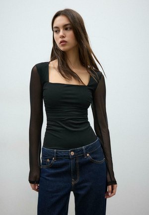 Giovane donna con indosso una maglietta nera aderente con maniche lunghe trasparenti e jeans blu scuro, in piedi davanti a uno sfondo chiaro e semplice.