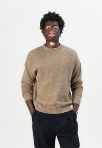 Maglione in maglia marrone con una finitura testurizzata, dotato di collo rotondo a coste e polsini. Abbinato a pantaloni scuri e larghi.