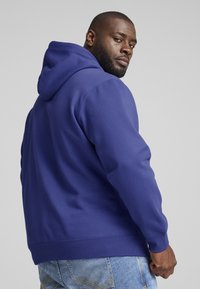 Felpa viola con cappuccio regolabile, polsini e orlo a costine. Presenta una texture liscia, indossata sopra jeans azzurri chiari. Design semplice e pulito.