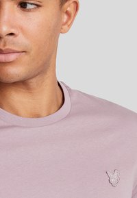 T-shirt di cotone lilla con scollatura rotonda e un piccolo logo di un uccello ricamato sul lato sinistro del petto. Texture liscia, vestibilità casual.
