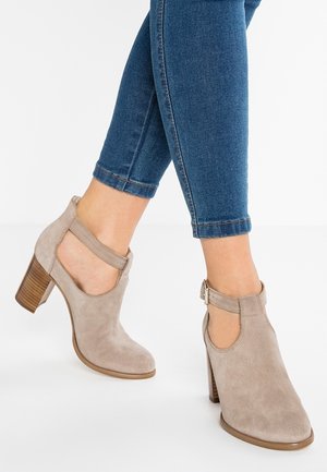 Beżowe zamszowe botki za kostkę z wycięciami, regulowanym paskiem i obcasem na słupku. Noszone z ciemnym deniem, prezentujące casualowy styl.