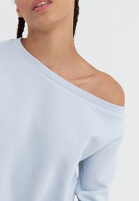 Kvinna med flätat hår som bär en ljusblå off-shoulder-topp, som visar hennes vänstra axel och en del av hennes nacke och ansikte.