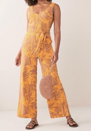 Femme portant une combinaison orange sans manches avec imprimé floral et ceinture nouée à la taille, tenant un sac à main rond tressé et portant des sandales noires.