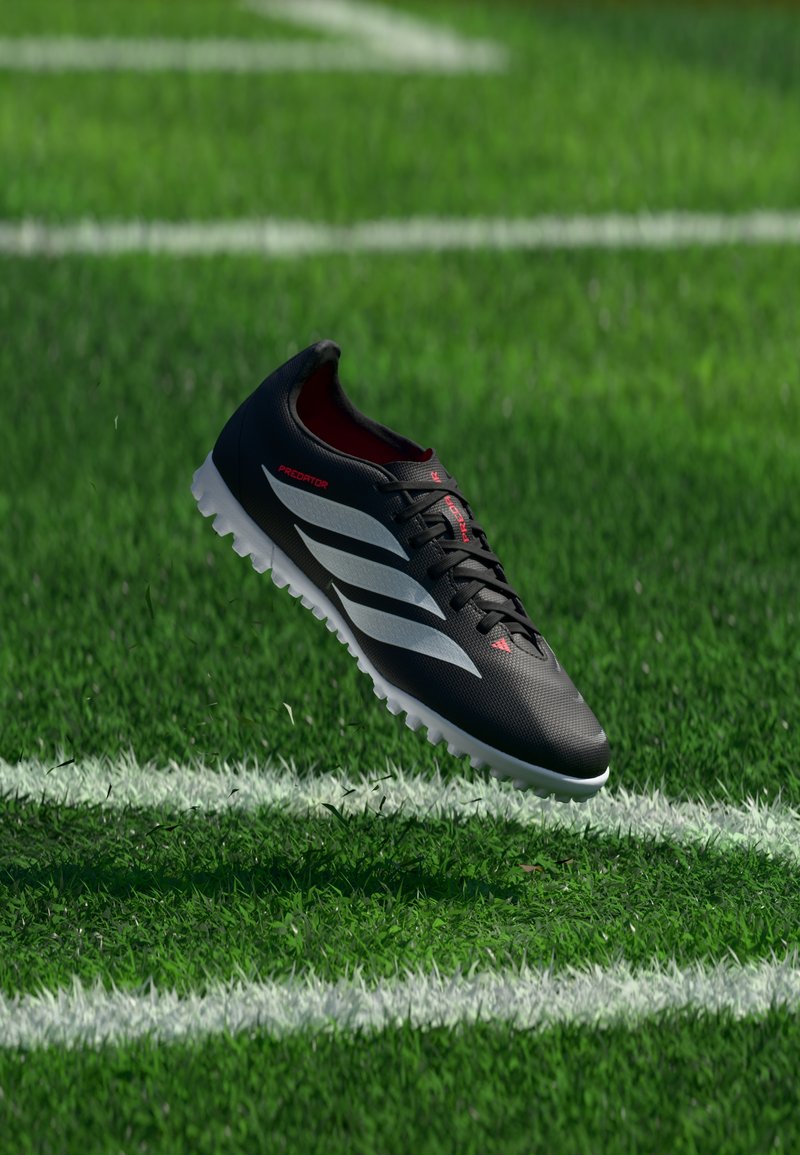 Chaussure de football noire avec des rayures blanches et des touches rouges flottant au-dessus d'un terrain de football vert avec des lignes blanches.