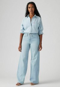 Lichtblauwe denimshirt met knoopsluiting, gecombineerd met lichtblauwe jeans met hoge taille en wijde pijpen. Model draagt neutrale sandalen.