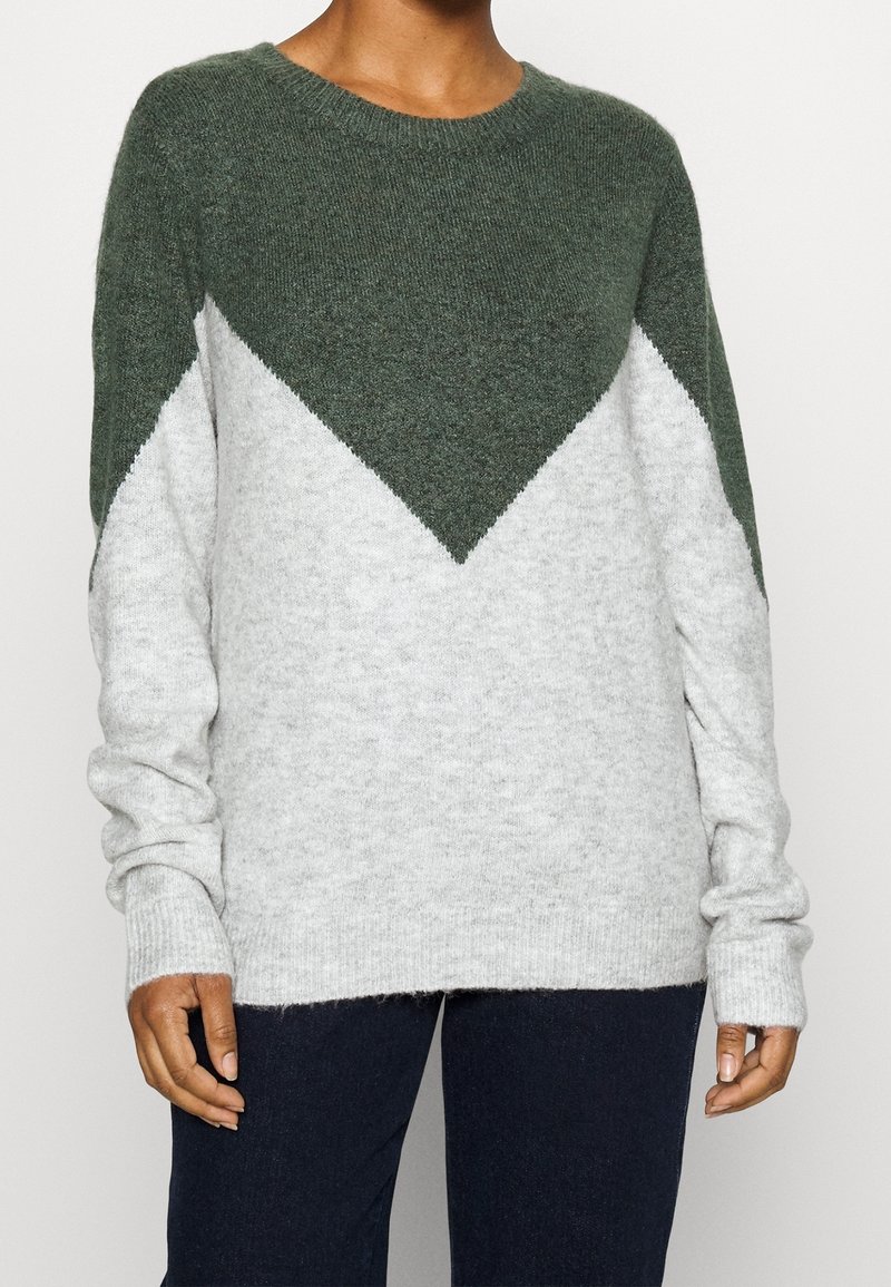 Personne portant un pull en maille à manches longues avec une partie supérieure vert foncé formant un triangle pointant vers le bas sur un corps gris clair.