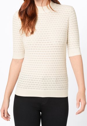 Vrouw in een crèmekleurige, kort mouw gebreide trui met een gevarieerd ruitpatroon en zwarte broek, zichtbaar van hals tot heupen.