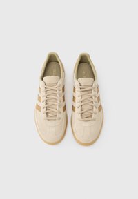 Beżowe zamszowe trampki z brązowymi paskami, zaokrąglonym noskiem, płaskimi sznurowadłami i gumowymi podeszwami w kolorze gumy. Wnętrze wyścielone jasnobeżowym materiałem z brandingiem.