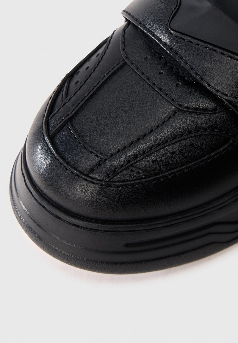 Nahaufnahme einer schwarzen Sneaker-Spitze aus Leder mit genähten Paneelen, perforierten Details und dicker Gummisohle auf weißem Hintergrund.