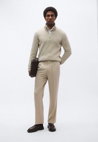 Beige strikket zip-up genser, kremfarget skjorte med krage, lys beige bukser, mørkebrune sko og en mørkebrun mappe. Ensfarget bakgrunn.