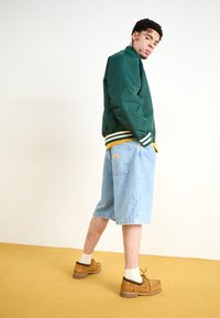 Jachetă bomber verde cu mâneci dungi, cămașă galbenă, pantaloni șort din denim albastru deschis și pantofi maro cu șosete albe, pe un fundal galben.
