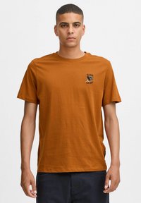 Brun bomulds T-shirt med rund hals, korte ærmer og et lille broderet "Born Lucky" logo på brystet. Glat tekstur.