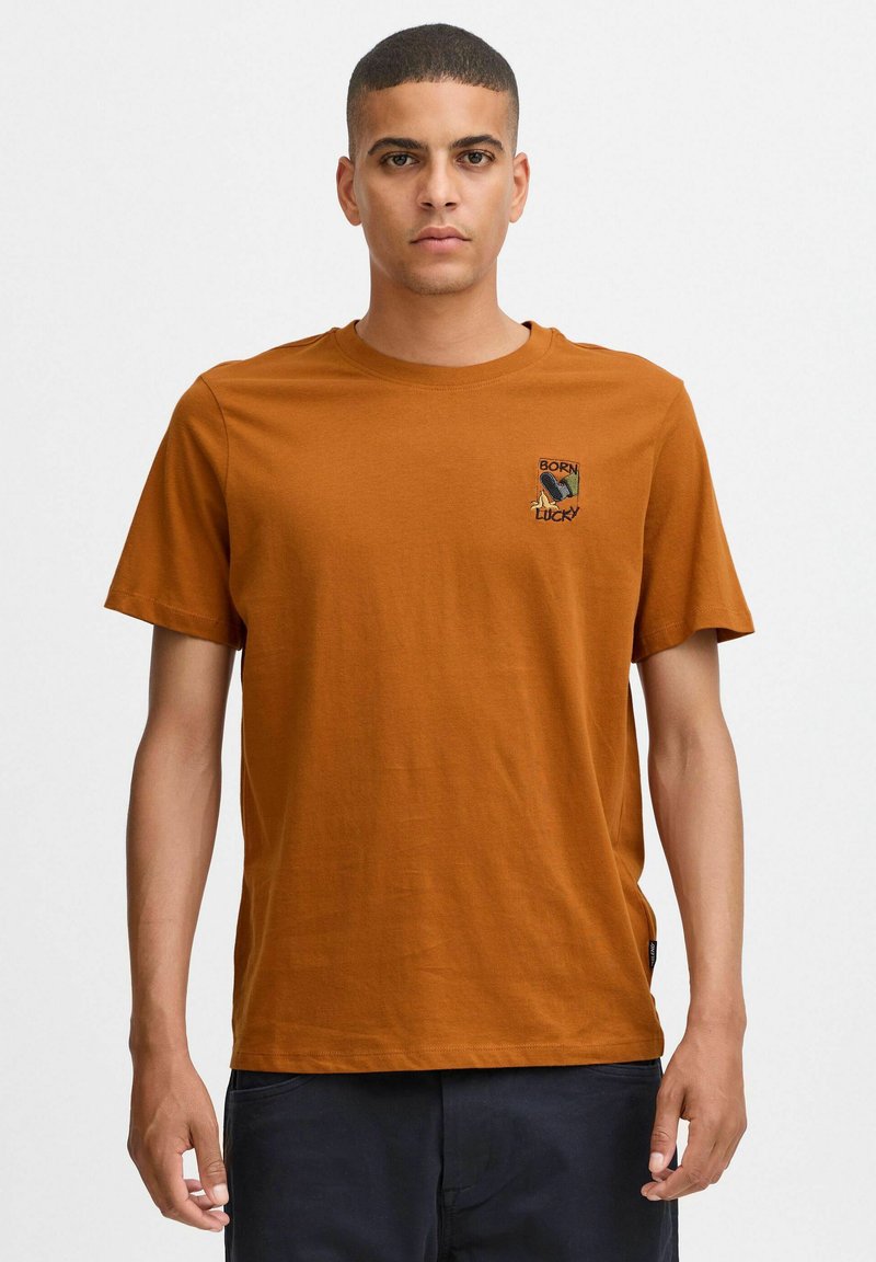 Brun bomulds T-shirt med rund hals, korte ærmer og et lille broderet "Born Lucky" logo på brystet. Glat tekstur.
