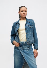 Τζιν μπουφάν μεσαίου ξεβγαλμένου χρώματος με κούμπωμα κουμπιών και τσέπες στο στήθος, συνδυασμένο με απαλό κίτρινο τοπ και τζιν φούστα με ασύμμετρο τελείωμα.