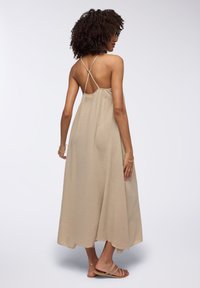 Abito maxi beige con spalline incrociate sottili, realizzato in un tessuto testurizzato e fluido che scende fino alle caviglie, abbinato a sandali piatti.