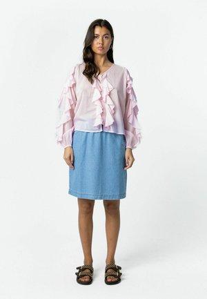 Žena oblečená v svetloružovej blúzke s volánmi, svetlomodrej rifľovej sukni po kolená a čiernych sandáloch s kovovými cvočkami stojaca na jednoduchom bielom pozadí.