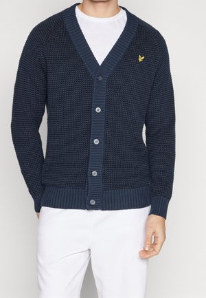 Mand iført en navy blå tekstureret cardigan med knapper med en gul fuglelogo på brystet og hvide bukser mod en ensfarvet baggrund.