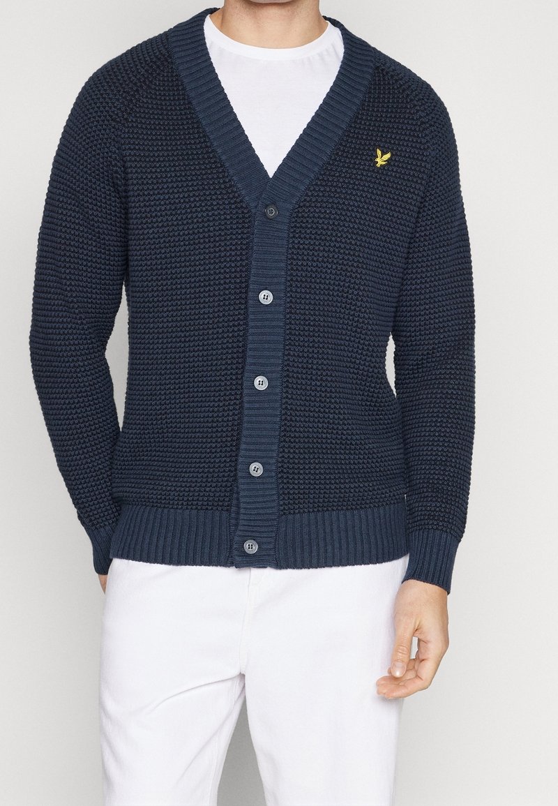 Homme portant un cardigan bleu marine texturé boutonné avec un logo d’oiseau jaune sur la poitrine et un pantalon blanc sur un fond uni.