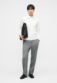 Άντρας που φοράει λευκό πόλο με μακριά μανίκια, γκρι παντελόνι με ρίγες, μαύρα παπούτσια, κρατώντας μεγάλη διπλωμένη μαύρη δερμάτινη τσάντα μπροστά σε λευκό φόντο.