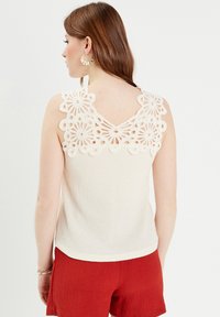 Haut sans manches de couleur crème avec un design en dentelle florale au niveau du col et un tissu texturé, associé à un short rouge.