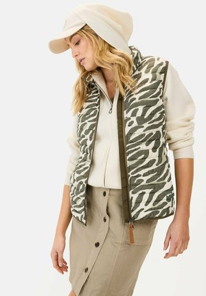 Grüne und cremefarbene Steppweste mit Zebra-Streifen, kombiniert mit einem hellen Reißverschluss-Sweater und einem beigen Rock mit Knopfdetails.