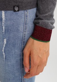 Pull gris avec poignets côtelés bordeaux et verts, tissu texturé, porté sur un jean bleu usé. Étiquette visible sur le côté du pull.