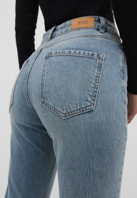 Světle modré denimové džíny s vyšším pasem, které mají výrazné zadní kapsy a hnědou koženou nášivku na pase. Texturovaná tkanina.